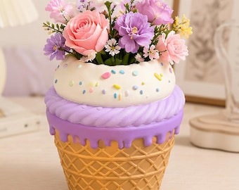 Könnte beinhalten: Ein dekorativer Übertopf in Form eines Eiswaffel-Cupcakes mit waffeltexturierter Basis, violettem Glasurguss und einer weißen Oberseite mit bunten Streuseln. Der Übertopf ist mit einem Arrangement aus künstlichen rosa, violetten und weißen Blumen gefüllt.