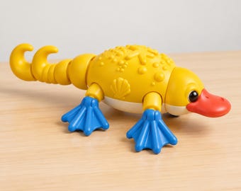 Könnte beinhalten: Ein Schnabeltier-Spielzeug aus gelbem Kunststoff mit einem leuchtend roten Schnabel und blauen Schwimmfüßen. Der Körper weist geformte Texturen auf, darunter kleine Noppen und ein Muscheldesign an der Seite. Der Schwanz ist segmentiert und gebogen.