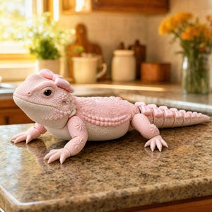 May include: A light pink articulated lizard figurine featuring a textured, scaly surface and a small bow accessory attached to the top of its head. The lizard has a rounded snout, visible eye detail, and a segmented tail extending behind its body.