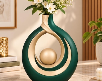 Könnte beinhalten: Eine kreisförmige Keramikvase mit einer tiefgrünen Außenseite und einer glatten, matten goldfarbenen Innenseite. Die Vase hat eine hohle Mitte mit einer schwebenden goldenen Kugel und enthält einen Strauß weißer Blumen mit grünen Blättern.