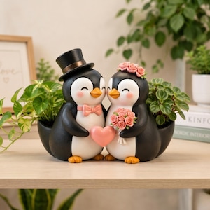 Puede incluir: Una maceta decorativa con dos figuras de pingüinos de pie uno al lado del otro. El pingüino de la izquierda lleva un sombrero de copa negro y una pajarita rosa, mientras que el de la derecha lleva una corona de flores rosas y un ramo.