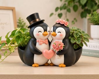 Könnte beinhalten: Ein dekorativer Übertopf mit zwei nebeneinander stehenden Pinguinfiguren. Der linke Pinguin trägt einen schwarzen Zylinder und eine rosa Fliege, während der rechte Pinguin einen rosa Blumenkranz trägt und einen Strauß rosa Rosen hält.