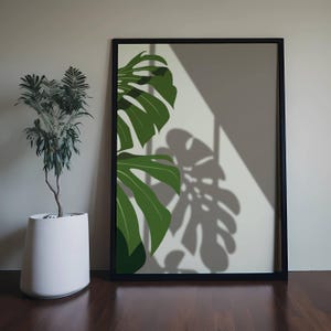 Könnte beinhalten: Ein gerahmter Kunstdruck mit einem grünen Monstera-Blatt-Design und einem schwarzen Rahmen. Das Kunstwerk wirft einen Schatten auf die Wand und erzeugt einen Kontrast zum Licht. Links steht ein weißer Pflanztopf mit einer grünen Pflanze.