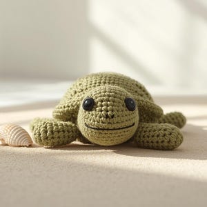May include: An olive green crocheted turtle with black eyes and a smiling face. A small seashell is next to the turtle. The turtle is on a light beige surface.