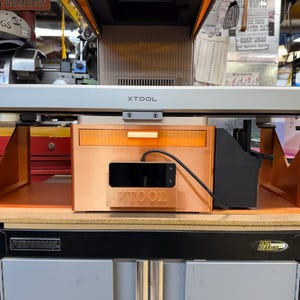 May include: A laser cutting machine with the brand name "XTOOL" visible. The machine has a gray frame and orange supports. A black screen is attached to the front. The machine is placed on top of a black and gray cabinet.
