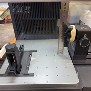 May include: A black and silver precision instrument with a ruler, a black box with a knob, and a black adjustable arm. The device is on a silver metal surface with a grid of holes.