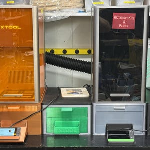 Peut inclure: Deux imprimantes 3D xTool avec des boîtiers rectangulaires hauts. L'unité de gauche a un couvercle orange transparent, tandis que celle de droite a un couvercle teinté foncé avec une étiquette rouge indiquant RC Short Kits & Prints.