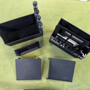 Puede incluir: Un surtido de componentes de plástico negro, posiblemente para un dispositivo electrónico o un sistema de almacenamiento modular, se disponen sobre una superficie verde. Las piezas incluyen cajas rectangulares, bandejas y una sección con elementos cilíndricos.