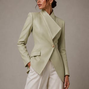 May include: A light green blazer with a unique asymmetrical design. The blazer features a high collar, a single gold button closure, and a small pocket. The model is wearing white pants. The blazer is made of a smooth, possibly leather-like material.