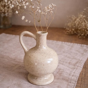 Op de afbeelding: Een gespikkelde, crèmekleurige keramische vaas met een handvat, gevuld met kleine witte bloemen. De vaas heeft een ronde vorm en een voet. De opstelling staat op een neutraal gekleurd linnen doek, wat een zachte, natuurlijke uitstraling creëert.