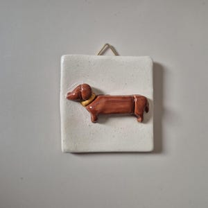 Op de afbeelding: Een keramieken teckel hond figuur in bruin met een gouden halsband, gemonteerd op een witte rechthoekige tegel. De tegel heeft een kleine lus om op te hangen. De hond is een gestileerd, decoratief stuk.