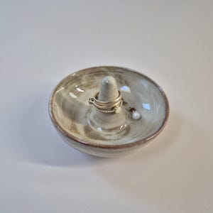 Op de afbeelding: Een keramische ringhouder met een centrale kegel en een ondiepe schaal. De houder heeft een gevlekte beige en bruine kleur, met een glanzende afwerking. Er liggen meerdere ringen en een kleine oorbel op de houder.