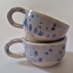 Op de afbeelding: Twee keramische mokken verticaal gestapeld, elk voorzien van een licht lavendelkleurig glazuur met donkerblauwe spikkelpatronen. De mokken hebben een ronde vorm en een enkel gebogen handvat aan de zijkant met een gladde afwerking.