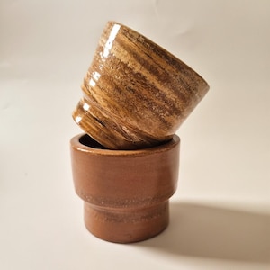 Op de afbeelding: Twee gestapelde keramische bekers. De onderste beker is effen, glad en mat terracotta bruin met een getrapte cilindrische vorm. De bovenste beker staat schuin en heeft een glanzende, gemarmerde afwerking met wervelende patronen.