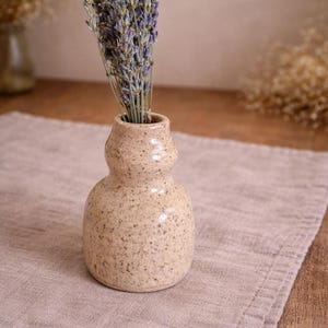 Può includere: Un piccolo vaso in ceramica beige macchiato contiene un mazzo di lavanda secca. Il vaso è appoggiato su un panno di lino dai toni neutri, con una superficie in legno in primo piano. La composizione evoca un'estetica rustica e naturale.