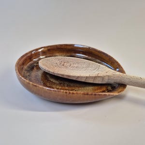 Op de afbeelding: Een bruine keramische lepelhouder met een houten lepel. De lepelhouder is ovaal en heeft een glanzende afwerking. De houten lepel is licht van kleur. Keukengerei.