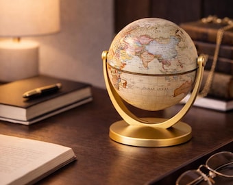 Op de afbeelding: Een vintage-stijl wereldbol met een crème-kleurige kaart, gemonteerd op een goudkleurige metalen standaard. De wereldbol, met het opschrift "Indian Ocean", staat op een donkerhouten bureau, naast een lamp, boeken en een dienblad met een bril.