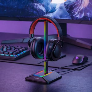 Puede incluir: Auriculares para juegos negros con detalles rojos sobre un soporte negro con una pantalla de luz LED arcoíris. Se ven un teclado y un ratón, lo que sugiere una configuración de juego. El soporte tiene el texto "Fast Store".