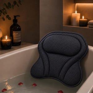 Può includere: Un cuscino da bagno grigio antracite scuro con una superficie testurizzata a rete e una forma ergonomica sagomata. Il cuscino è posizionato contro il bordo interno di una vasca da bagno bianca riempita d'acqua e petali di rosa rossi.