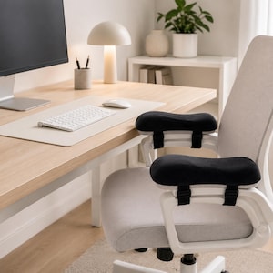 May include: A pair of black, contoured armrest pads attached to the arms of a light gray office chair. The pads feature a soft, textured surface and are secured to the chair frame with adjustable black straps, providing cushioned support.