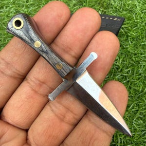 Könnte beinhalten: Ein Miniaturdolch mit polierter Stahlklinge und dunklem Holzgriff. Der Griff ist mit Messingnieten und einem messingfarbenen Ring verziert. Der Dolch hat einen Parierstange und eine Spitze.