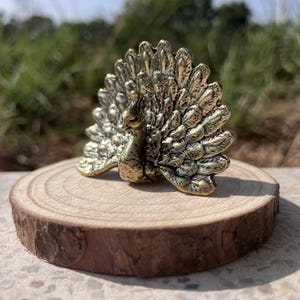 May include: A detailed brass peacock figurine with its tail feathers fanned out, displayed on a small, round wooden slice. The peacock is a golden color, and the wood slice has a natural, rustic appearance.