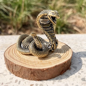 May include: A detailed brass cobra figurine with an open mouth, coiled on a small wooden slice. The snake's scales and features are intricately designed, showcasing a vintage aesthetic. The cobra is a golden color.
