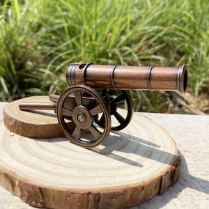 May include: A small bronze cannon model with spoked wheels and a detailed barrel. The cannon is displayed on a wooden slice, showcasing its antique design. The background features green grass.