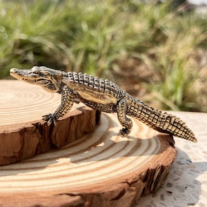 May include: A bronze-coloured crocodile figurine with detailed scales and legs, positioned on a natural wood slice. The figurine has an open mouth and is set against a blurred green background.
