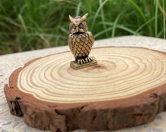 May include: A small, detailed brass owl figurine with textured feathers and large eyes. The owl is perched on a small rectangular base, set on a round, natural wood slice with visible growth rings. The background is blurred green foliage.