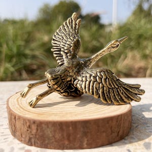 Op de afbeelding: Een bronzen metalen sculptuur van een vogel in vlucht, met uitgespreide vleugels, gemonteerd op een kleine, ronde houten basis. De veren van de vogel zijn gedetailleerd en de sculptuur heeft een antieke afwerking. De houten basis heeft een natuurlijke, rustieke uitstraling.