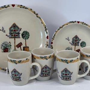 May include: A dinnerware set featuring two cream-colored ceramic plates and four matching mugs. Each piece is decorated with a painted blue birdhouse, small birds, and a green topiary, framed by a yellow border with green leaves and red hearts.