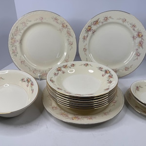 May include: A collection of cream-colored vintage dinnerware with floral accents. The set includes large plates, bowls, and smaller dishes, all with a gold trim. The floral pattern features pink and yellow flowers.