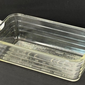 May include: A clear, rectangular glass baking dish with ribbed sides and decorative handles. The dish is empty and sits on a dark surface. This vintage-style dish is suitable for baking and serving.