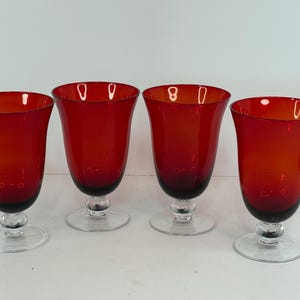 May include: A set of four matching drinking glasses featuring deep red, translucent bowls that taper toward the base. Each glass is supported by a clear, circular pedestal foot and a short, rounded stem, creating a uniform and elegant silhouette.