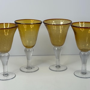 May include: Four amber-colored wine glasses with clear stems and bases. The glasses have a textured appearance and a flared bowl shape. The stems are bulbous and the bases are round. The glasses are arranged in a row.