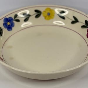 May include: Off-white ceramic bowl with a floral design. The rim features a garland of flowers in blue, yellow, and maroon, with green leaves. A thin maroon line borders the inside edge of the rim. The bowl has a slightly worn appearance.