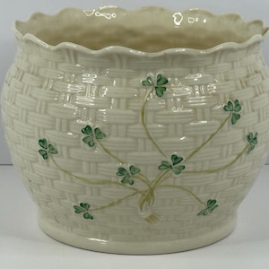 May include: Cream-colored ceramic planter with a woven basket design. The planter features green shamrock accents and a scalloped rim. The round planter is suitable for indoor or outdoor use.