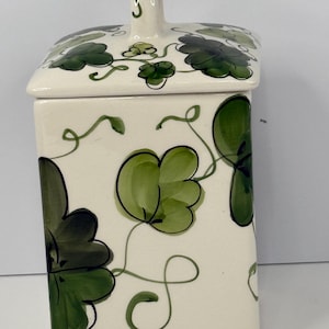 May include: A rectangular, off-white ceramic jar with a lid, adorned with green floral designs. The jar features a handle on the lid and is decorated with stylized green leaves and vines. The design is hand-painted.
