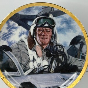 May include: A round decorative collector plate featuring a detailed illustration of a pilot in a cockpit, wearing a white helmet with goggles and a flight suit. The plate has a gold-toned rim decorated with small star motifs along the edge.