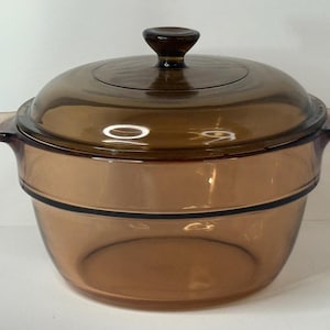 May include: A transparent amber-colored glass casserole dish with a matching lid. The dish has two side handles and a dark brown knob on the lid. The cookware is designed for oven use.