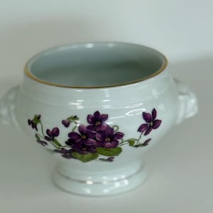 Puede incluir: Cuenco de cerámica blanca con borde dorado y dos asas pequeñas. El cuenco está decorado con flores de violetas moradas y hojas verdes. El cuenco se asienta sobre una pequeña base con una banda plateada.