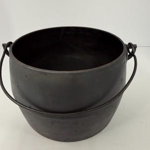 May include: A dark charcoal-colored cast iron pot with a rounded body and a wide, open top. The pot features a thin, curved metal handle attached to two side lugs, allowing it to swing freely. The exterior surface has a matte, textured finish.