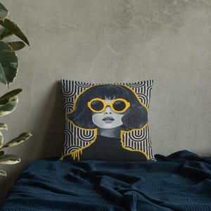 May include: Square throw pillow featuring a stylized portrait of a woman with dark hair and yellow sunglasses. The pillow has a black and white geometric pattern background with yellow accents. The woman is wearing a black turtleneck.