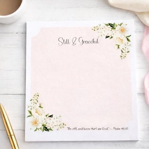 May include: A light pink notepad with floral corner designs and the text "Still & Graceful." The notepad features a quote from Psalm 46:10. A gold pen and a cup of coffee are visible in the background.