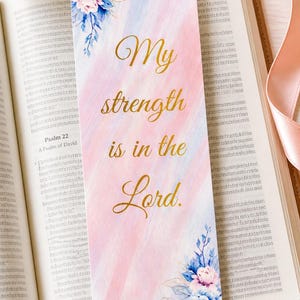Könnte beinhalten: Ein Lesezeichen mit dem Text "My strength is in the Lord" in goldener Schrift. Das Lesezeichen hat einen rosa-blauen Aquarellhintergrund und florale Akzente. Es liegt in einem offenen Buch.