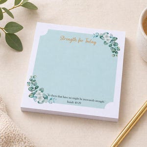May include: A square notepad with a light blue background and floral accents. The text "Strength for Today" is in gold script. The notepad is next to a gold pen and a cup of coffee.