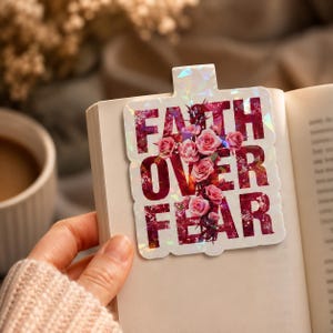 Könnte beinhalten: Ein weißes Lesezeichen mit den Worten "FAITH OVER FEAR" in Burgunderrot, mit einem Bouquet aus rosa Rosen. Das Lesezeichen hat ein holografisches, irisierendes Finish und befindet sich in einem offenen Buch.
