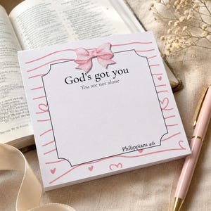 May include: A square notepad featuring a pink watercolor bow at the top, pink wavy lines, and small hearts. The text reads God's got you, You are not alone, and Philippians 4:6. The notepad has a white center area with a black decorative border.