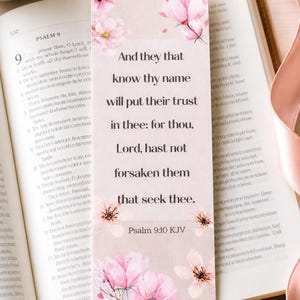 May include: A bookmark with a quote from Psalm 9:10 KJV, decorated with pink flowers. The bookmark is placed on an open book, with a gold pen and a pink ribbon.